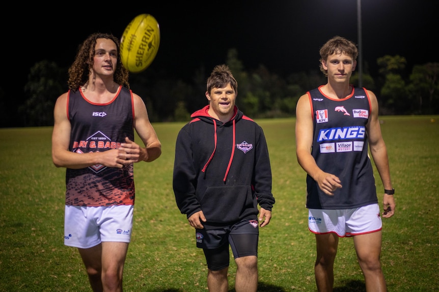 Three young men in sports gear walk off the field, while one tosses a football in the air
