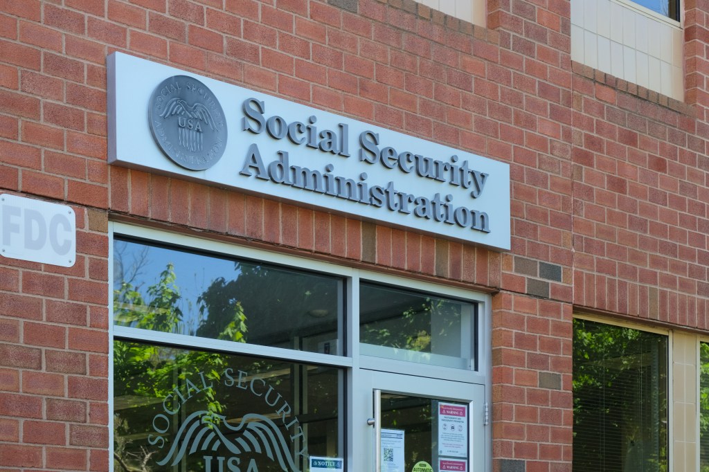 The sign for the Social Security Administration on a brick building, with a reflection of trees in the window.