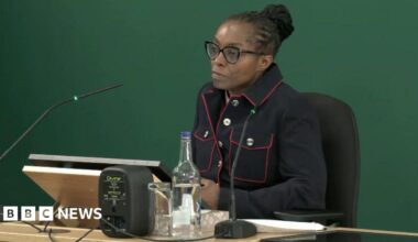 A woman sat a desk in a blue jacket with red trim in front of a green back drop.