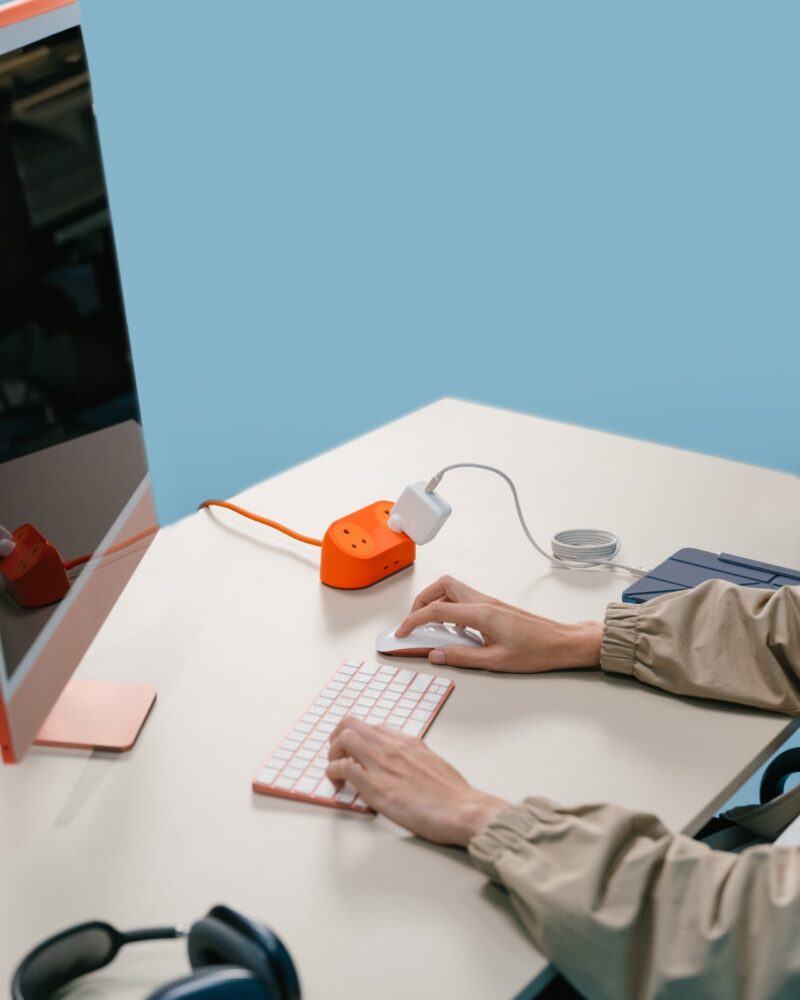 Person using a wireless mouse and keyboard at a desk with a desktop computer, DailyObjects orange power hub, and headphones on a light surface against a blue background.