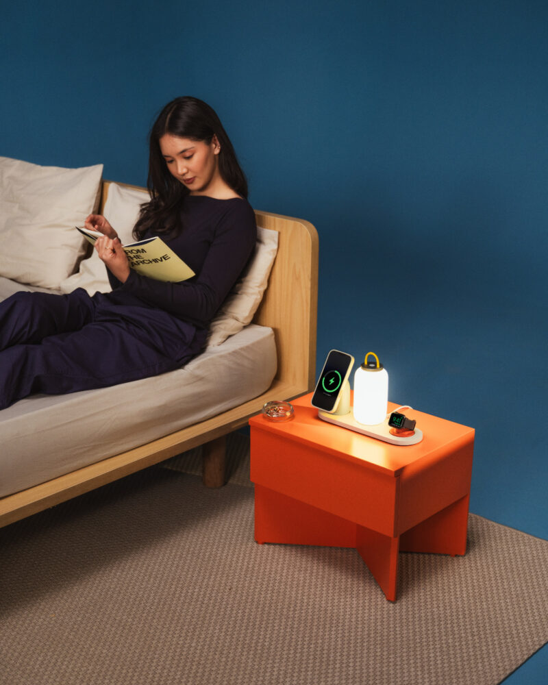 A woman sits on a bed writing in a notebook. Next to her is an orange DailyObjects nightstand with a lamp, a smartphone, earbuds, and a glass dish.