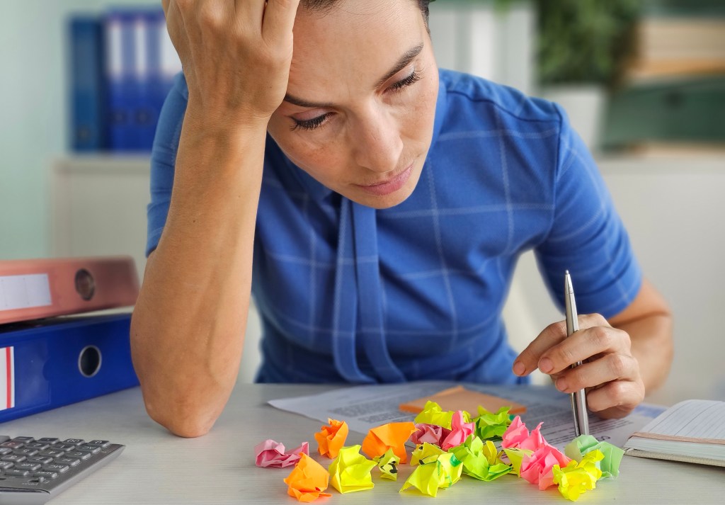 Woman at a desk, looking stressed, surrounded by crumpled colorful papers while working.