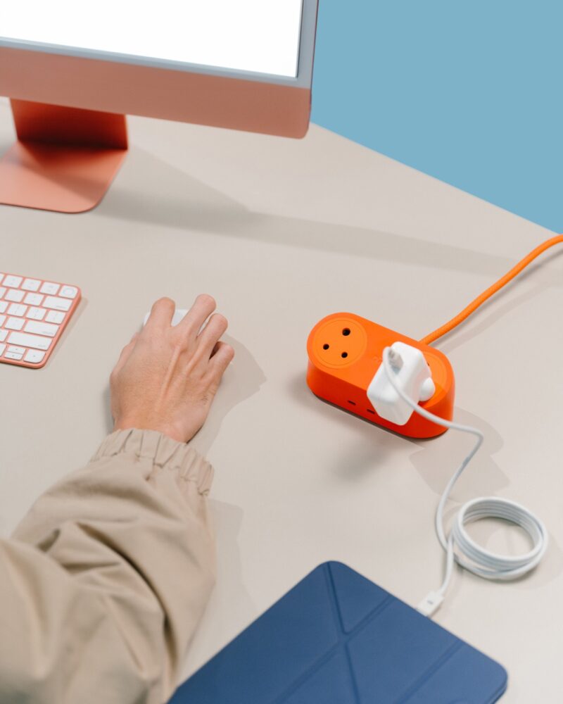 A person uses a desktop computer beside a DailyObjects orange power strip with a charging adapter and cable on a light-colored desk.