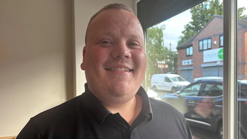 A jolly looking man in a black polo shirt and with short cropped hair smiles at the camera.