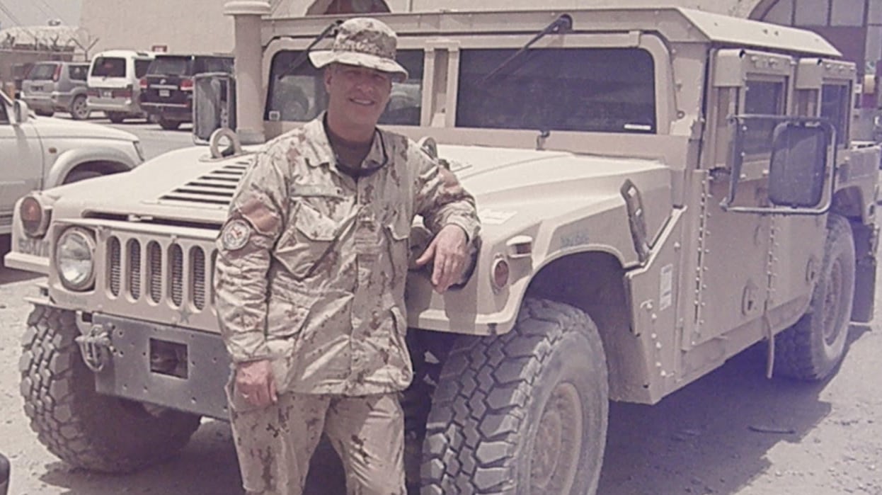A man in his fifties stands by a military vehicle wearing a camouflage military uniform.