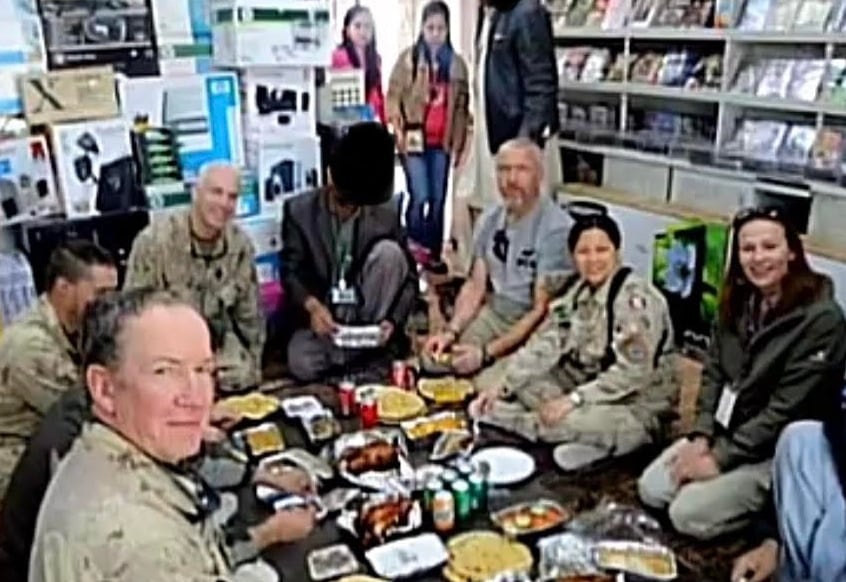 A group of men and women sit in a circle around a meal in a shop at the Kandahar Airfield in Afganistan. Some of them are there representing NATO countries that sent members on the ISAF mission.