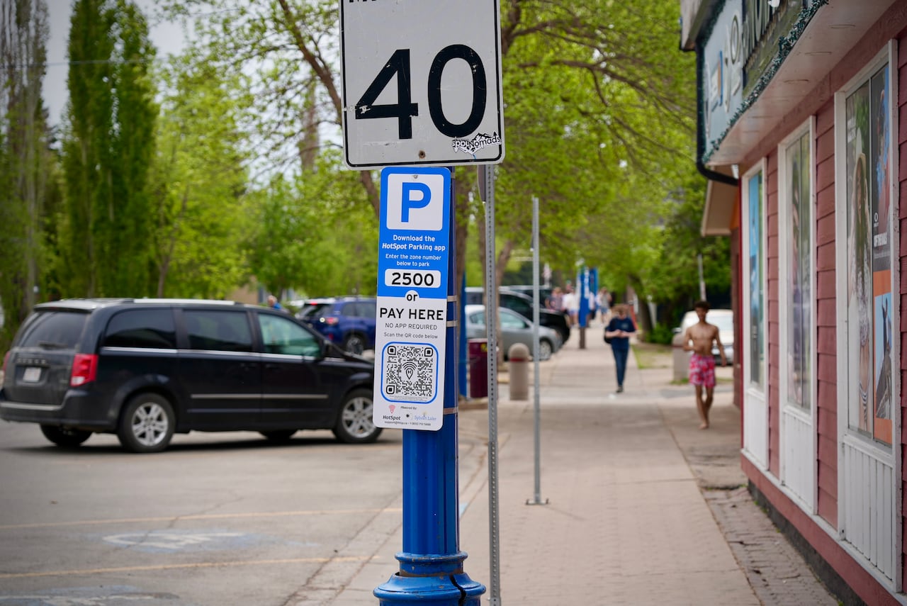 A parking sign tells people how to pay. Behind it, there are cards parked in front of the curb, while people walk along a sidewalk.