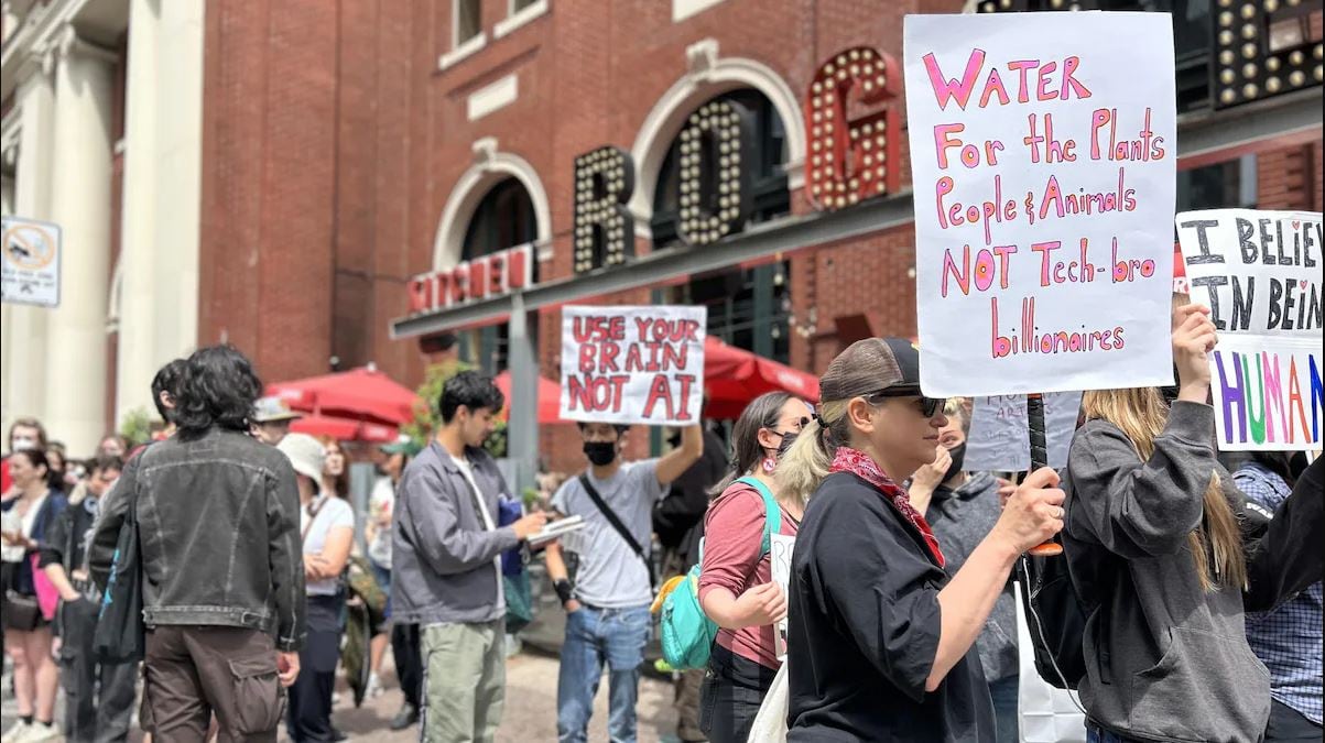 People walk outside a brick building holding protest signs, including one that says, “Water for the plants, people & animals, not tech-bro billionaires.”