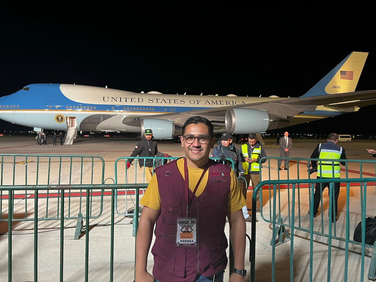 A man wiht glasses and press passes hanging from his neck stands by a metal fencing in front of the U.S. presidential plane Air Force 1.