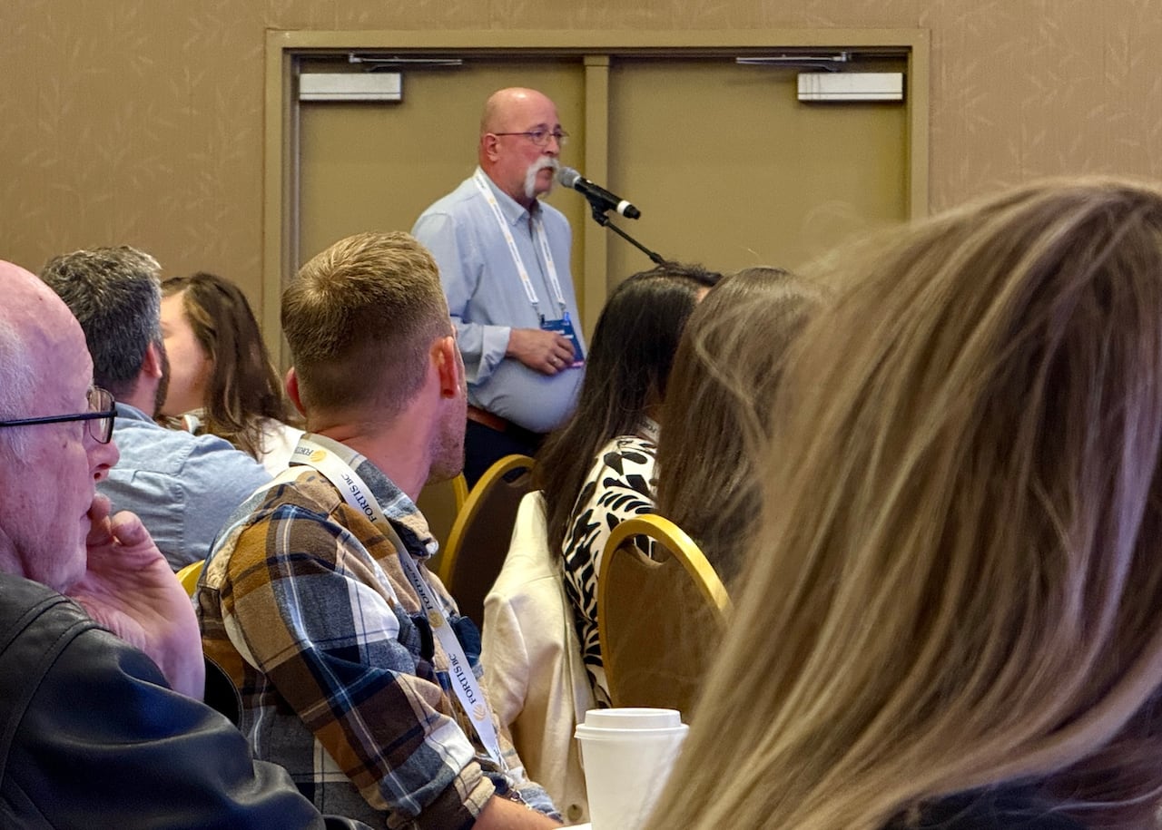 a man speaks into a microphone at the front of a conference room while attendees listen from their seats
