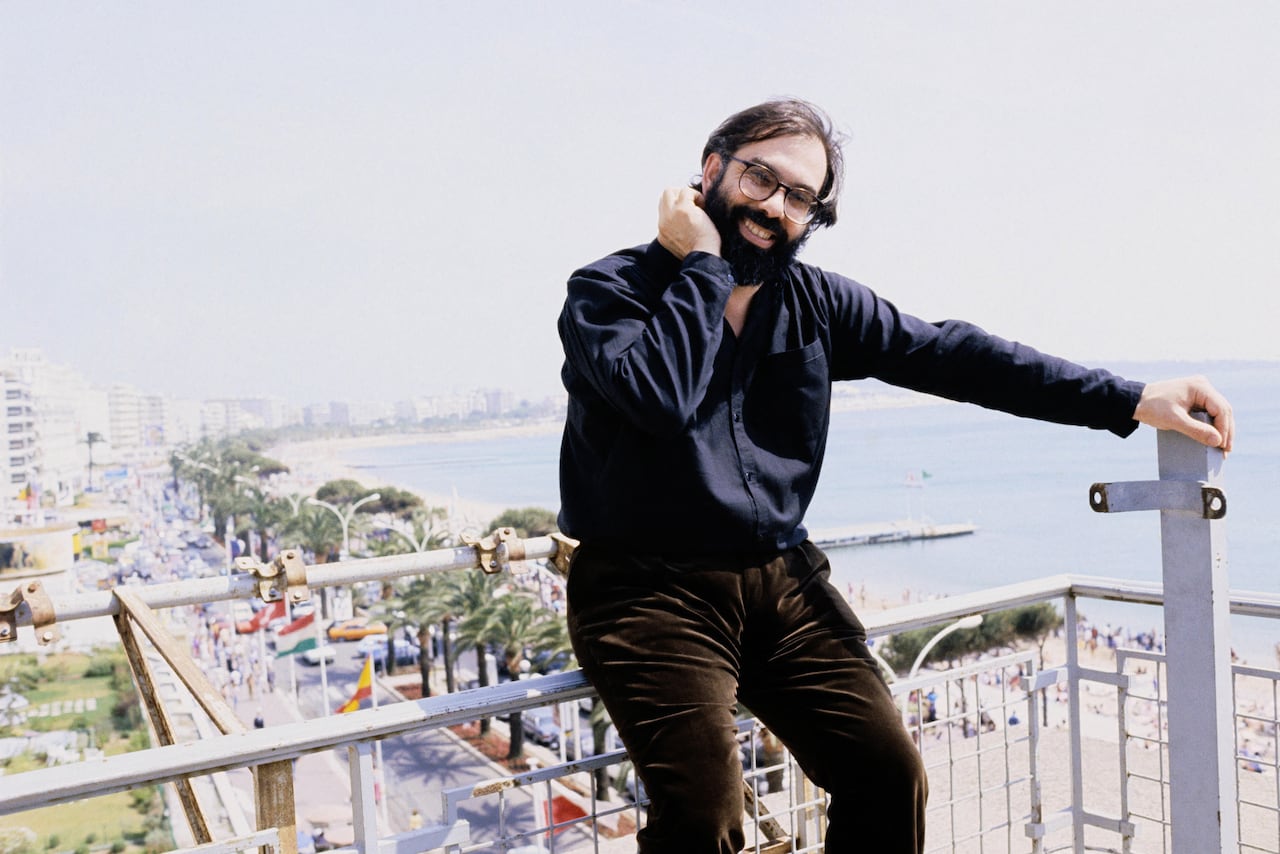A smiling bearded man in a dark top and pants sits on railing outdoors, with a beach and palm trees seen behind him.