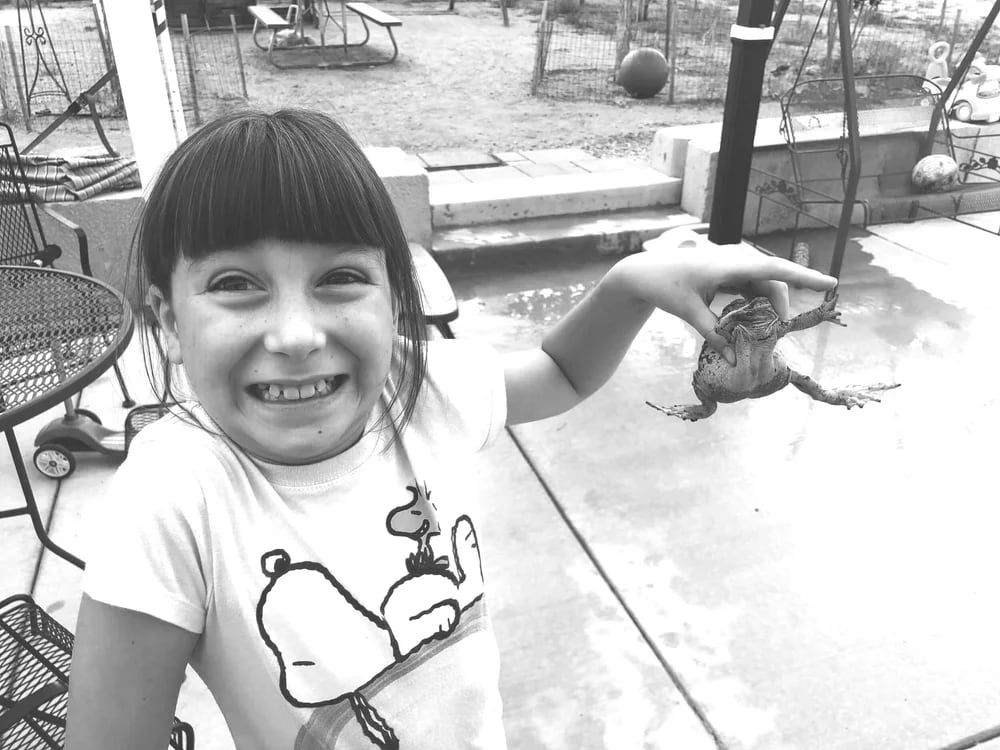 A girl holding up a frog by the tips of her fingers smiles and shrugs.