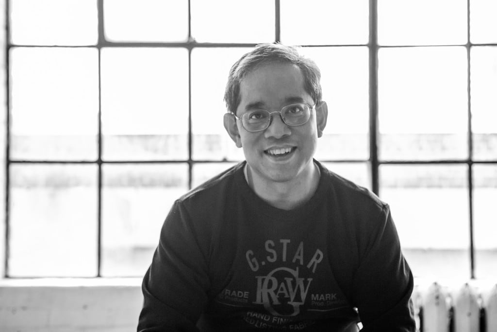 A black-and-white photograph shows a smiling man with glasses sitting in front of a window.