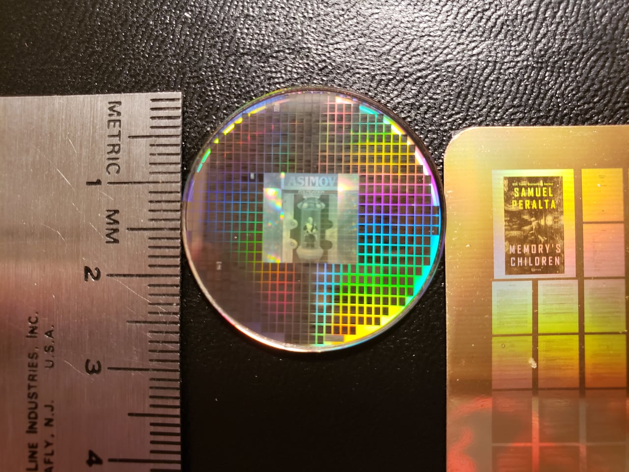 A small disc with dozens of tiny black squares rests next to a ruler on a black surface.