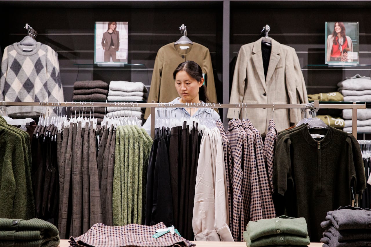 a woman with dark hair stands behind a rack of clothes, looking through the items