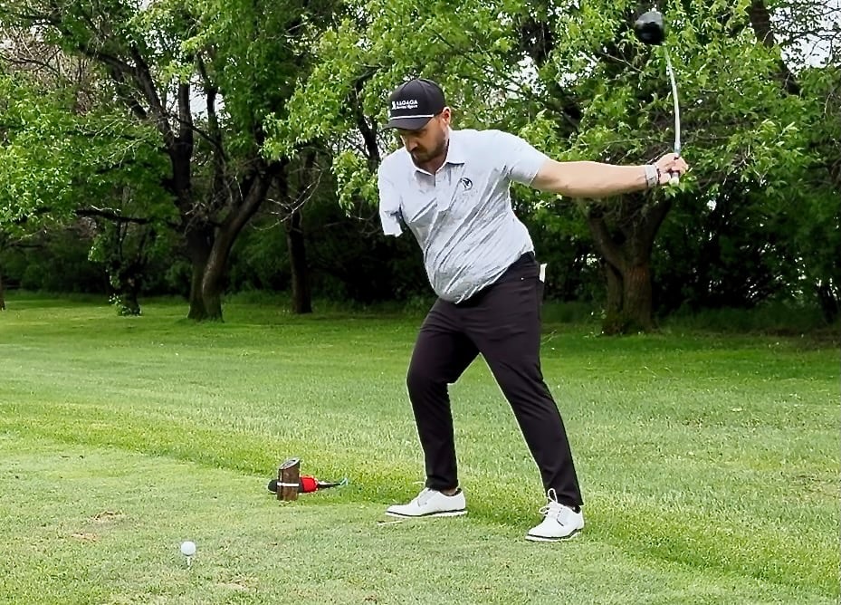 A driver appears to bend in a downward swing as a one-armed man in golf shirt cap and black pants drives the ball on a green tee-box of a golf course.