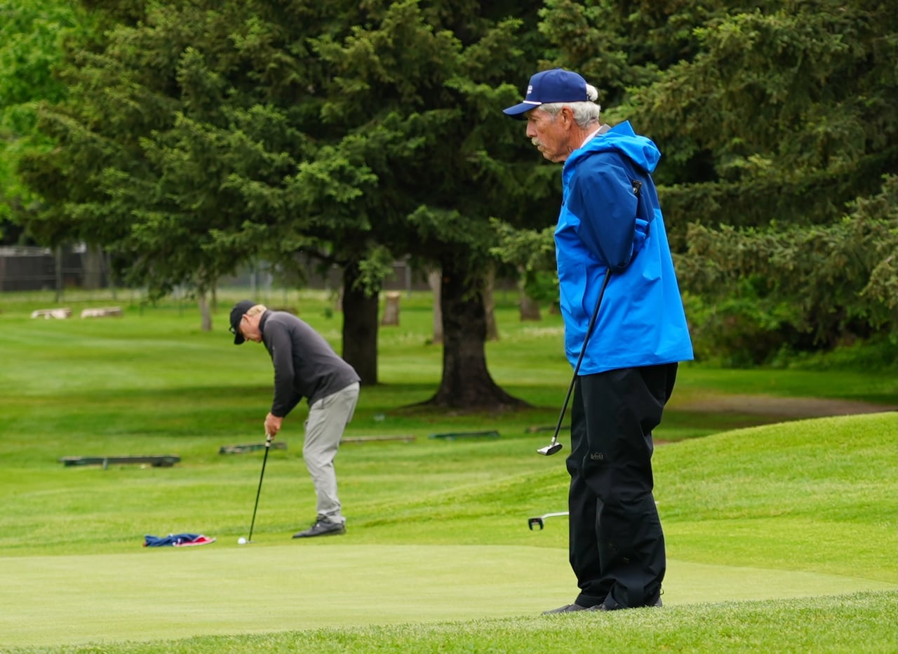 An older man in a blue windbreaker studies the putting green while holding a putter under his sleeve.