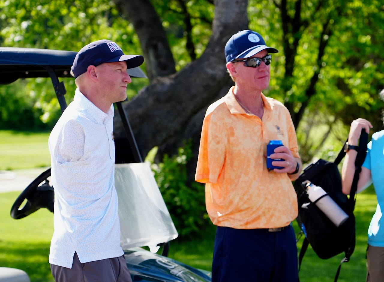 Two men in blue caps and golf attire lean on a golf cart and relax after a round of golf.