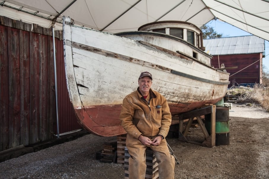 A man in work clothes sits in front of an old wooden fishing boat.