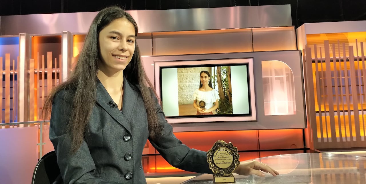 A woman sits at a table with an award in front of her and a photo on a screen behind her.