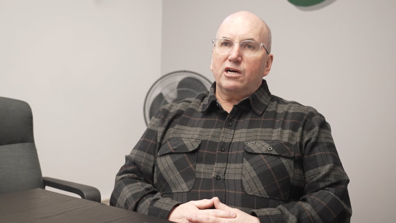 A man in a flannel shirt speaks while seated in a room.