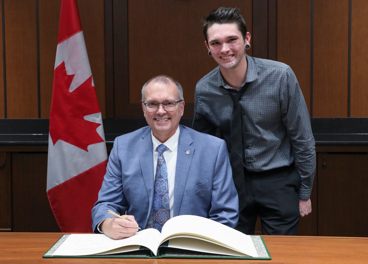 A man sits at a desk and a younger male stands beside him.