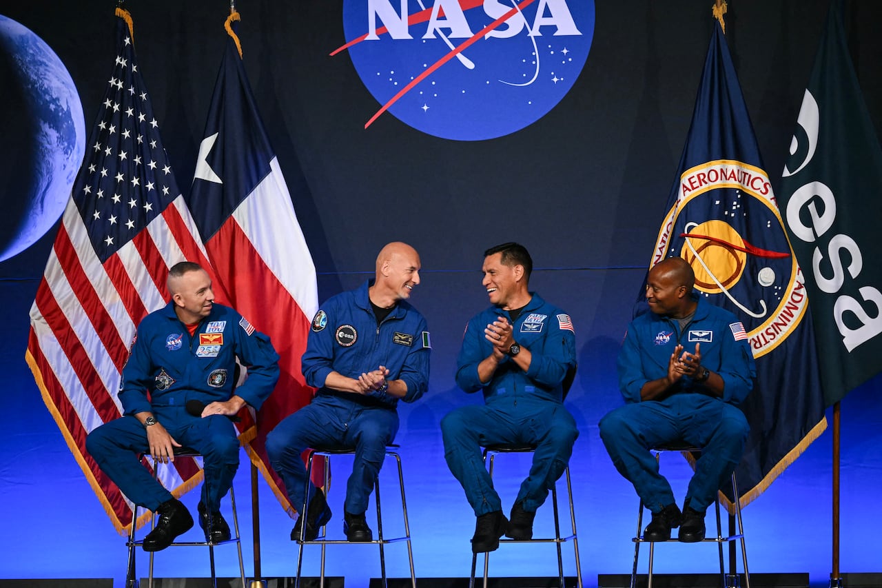 Four astronauts sit in blue jumpsuits in front of a large NASA logo on stage.