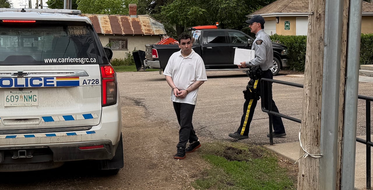 A man in a white shirt and handcuffs walks toward an RCMP cruiser.