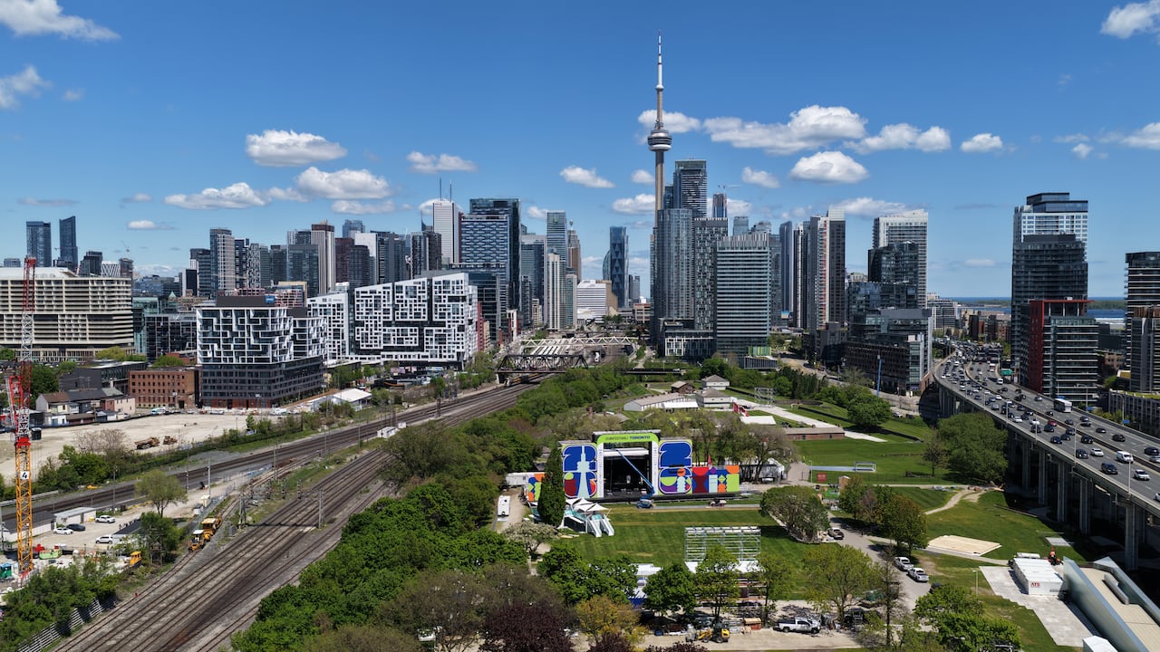 Aerial (drone) images of Fort York as it undergoes transformation to the FIFA World Cup Fan Festival. There is a stage with a sign for the fan festival, next to a highway, with the Toronto skyline and east end in the background. It is sunny.