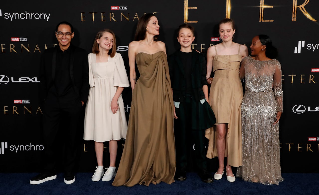 A woman stands in front of a movie backdrop with 2 children on her left and 3 children on her right. All are wearing formal attire.