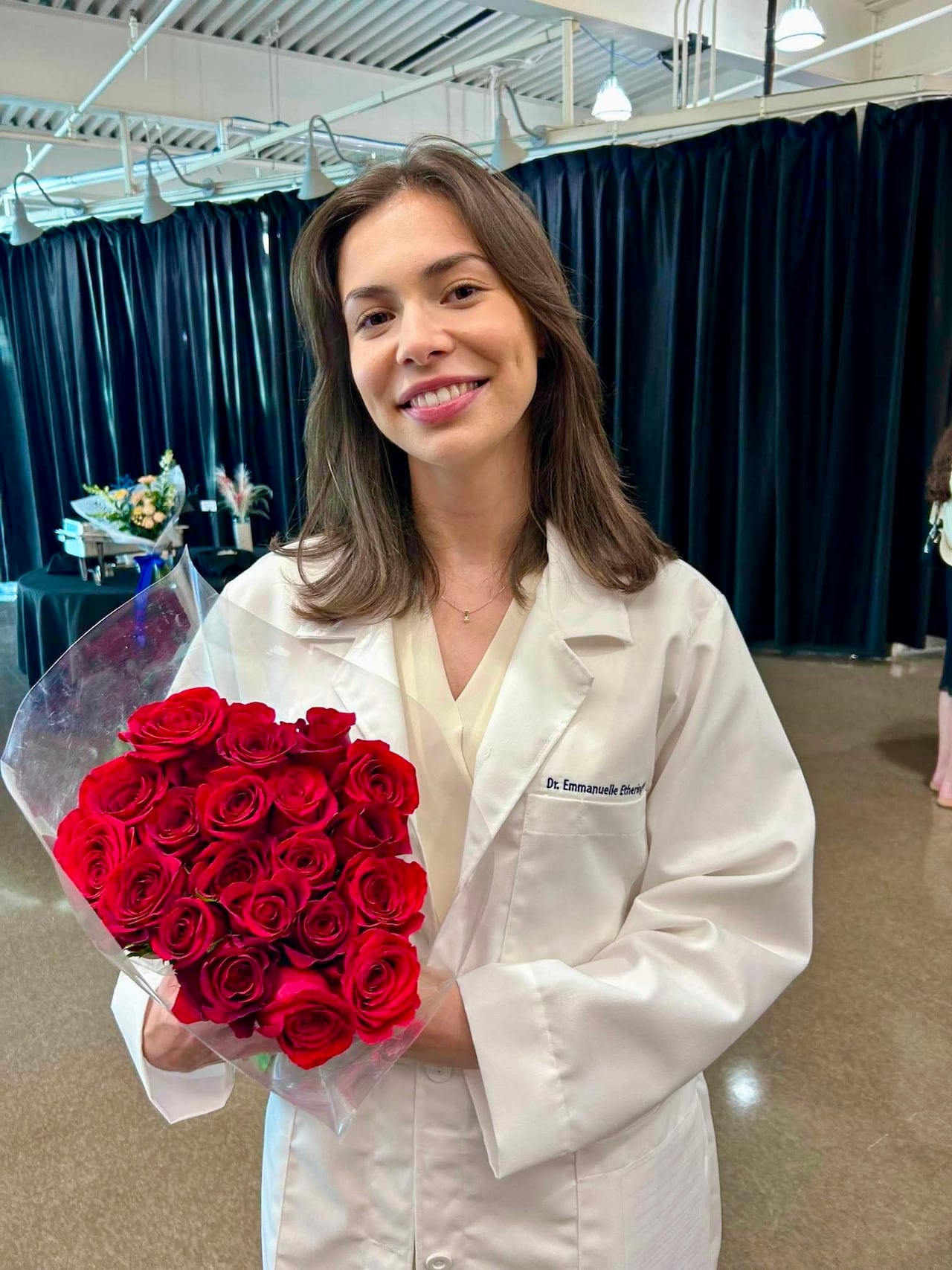 A young woman in a white doctor's coat holds a bouquet of red roses.