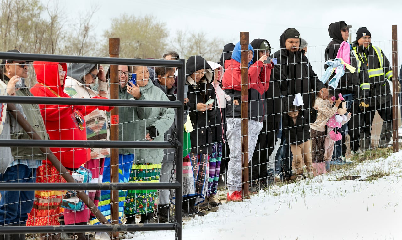 Women in skirts, children and men all stand behind a wired fence that is taller than a man's height.