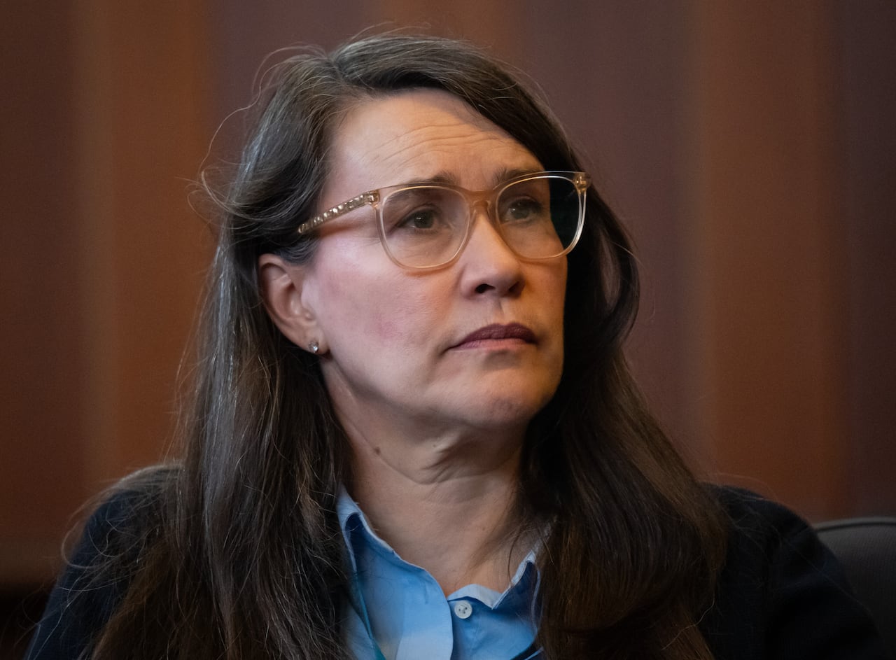 A white woman wearing glasses looks on.