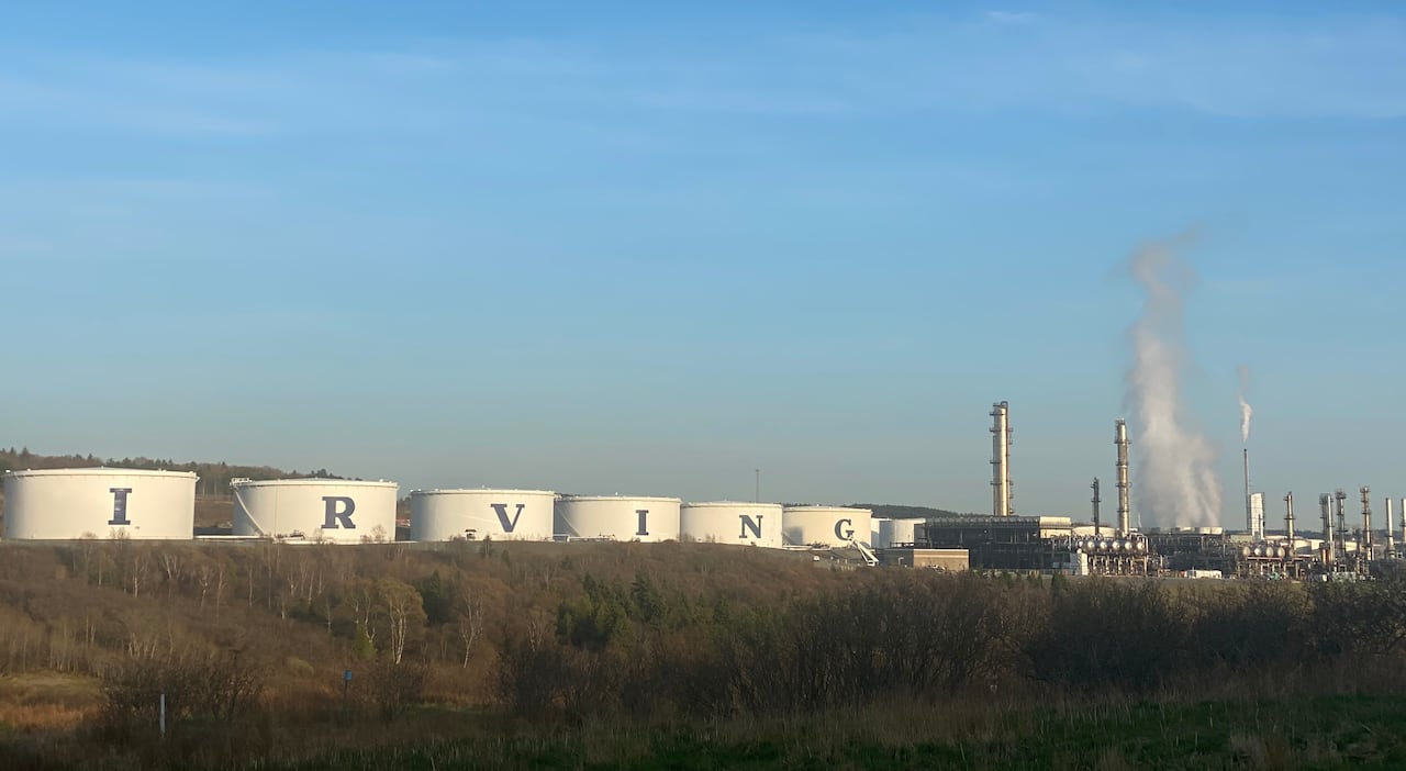 Six large oil tanks beside a refinery with smoke billowing from its stacks.