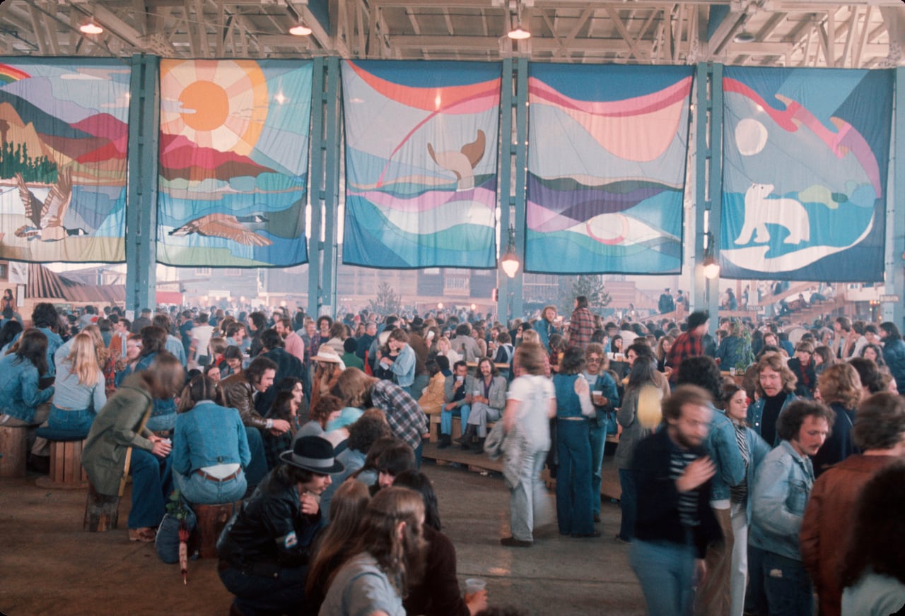 A vintage 1970s photo of large batik banners hanging in a refurbished airplane hangar as throngs of people congregate and socialize.