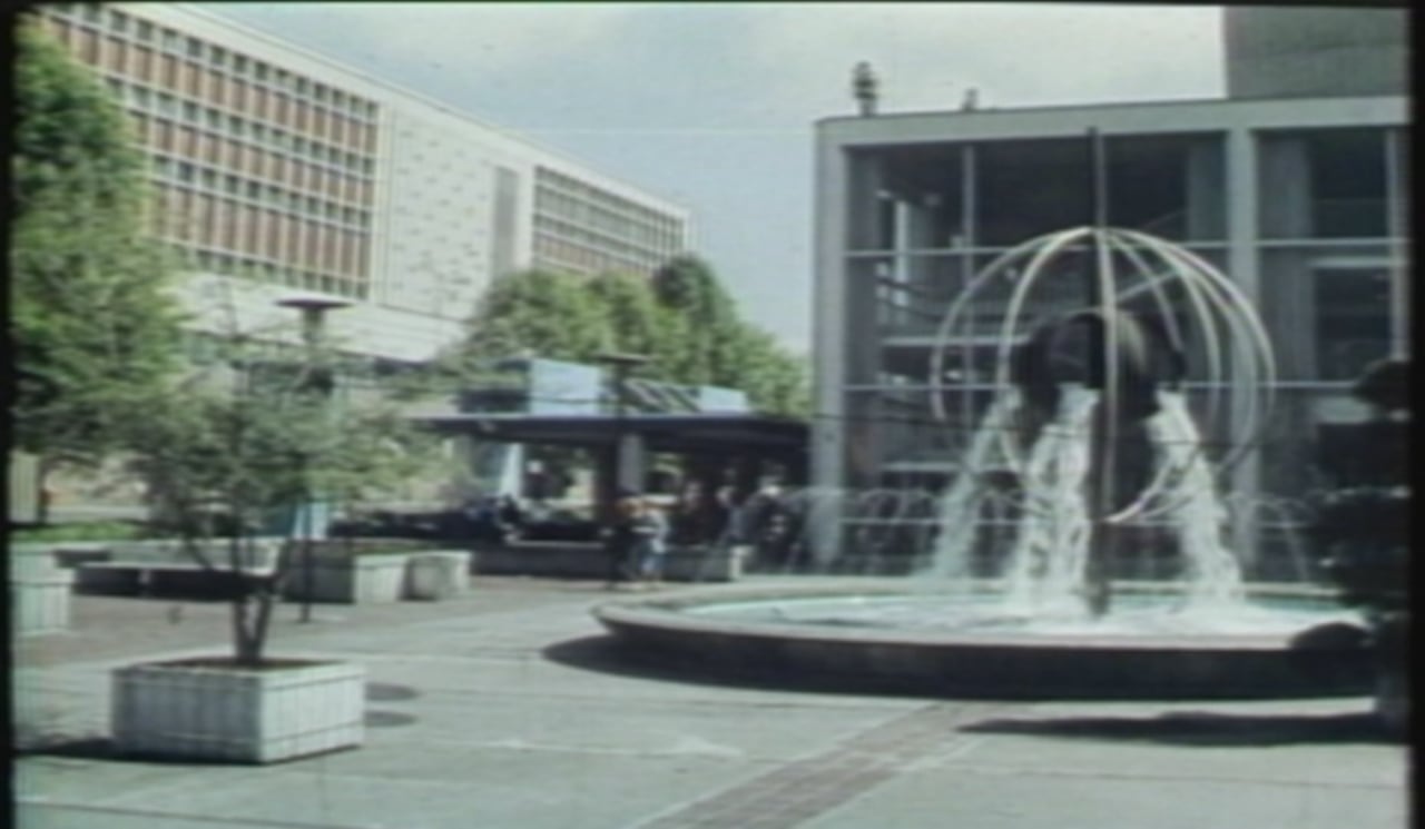 A vintage still photo of a plaza with a fountain and several buildings.