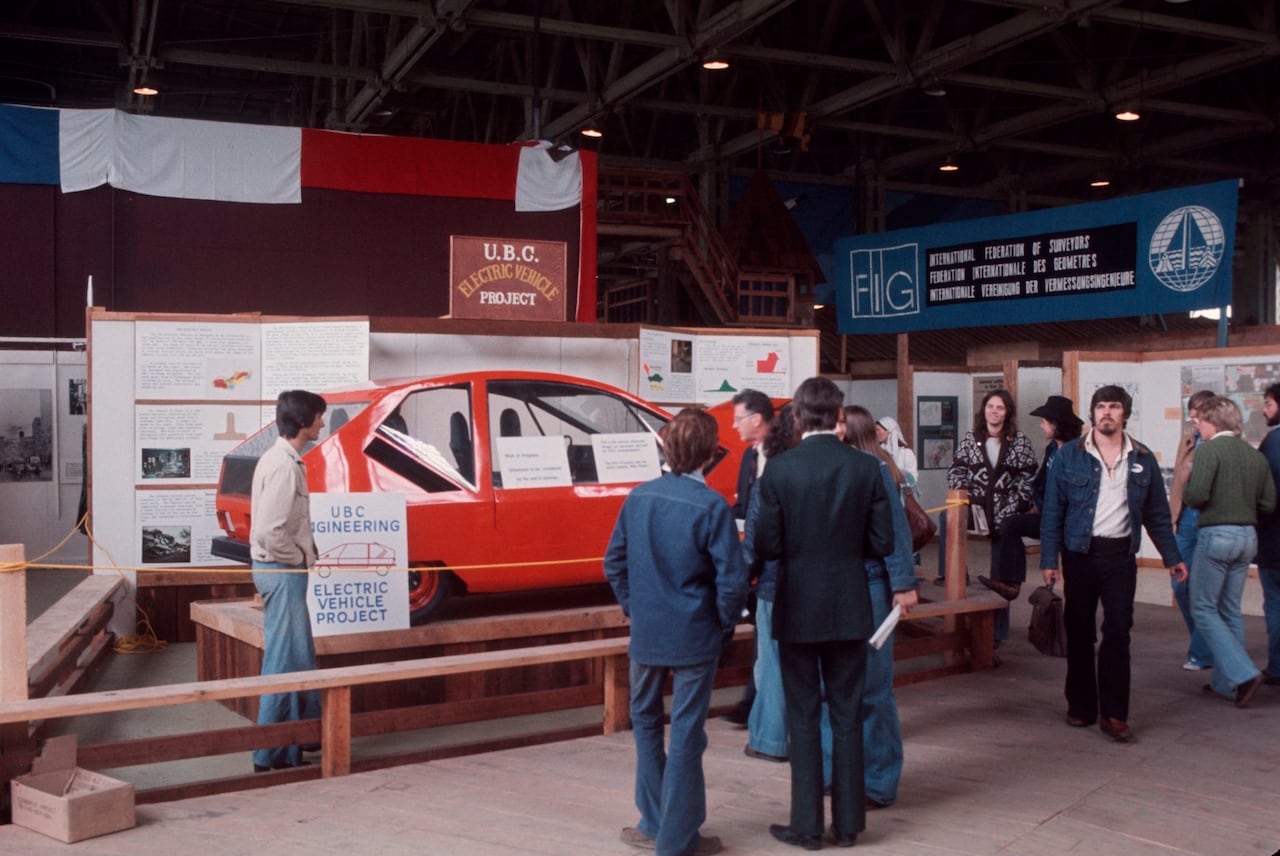 A vintage photograph showing a red car with a sign indicating it is a 'UBC Engineering: Electric Vehicle Project.' People gather around the cordoned off car, as a person behind the rope looks at the coupe-style car.