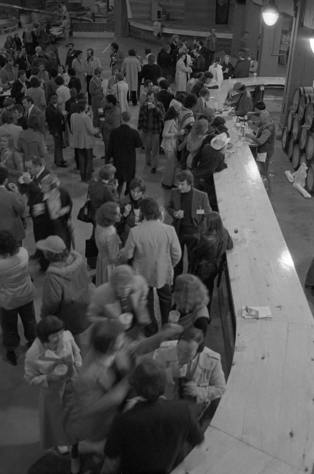 A black and white vintage photo showing people congregating at a very long wooden bar.