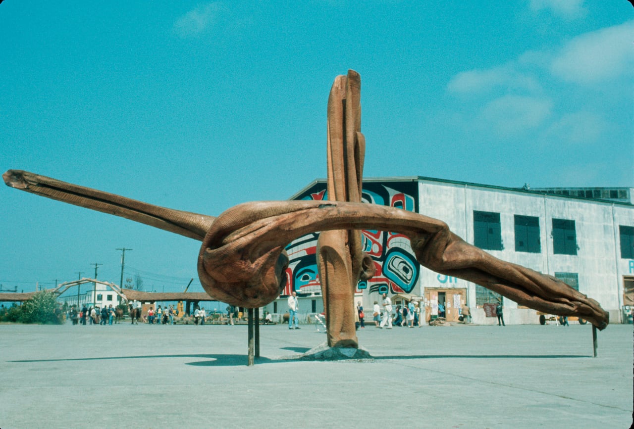 A large wooden sculpture stands in a prominent position at the Habitat Forum at Jericho Beach near an airplane hangar under blue skies.