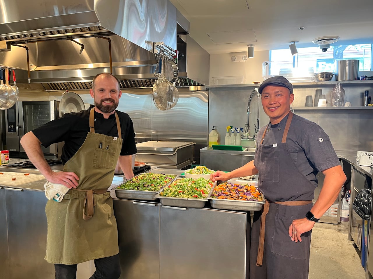 Two men wearing aprons in a kitchen with trays of food