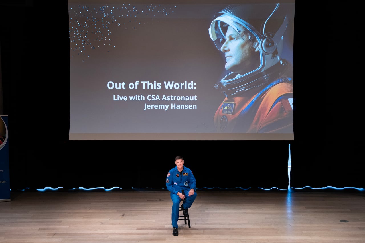 A man in a bright blue jumpsuit sits on a chair on a stage with an image projected behind him that says "Out of this World: Live with CSA Astronaut Jeremy Hansen."