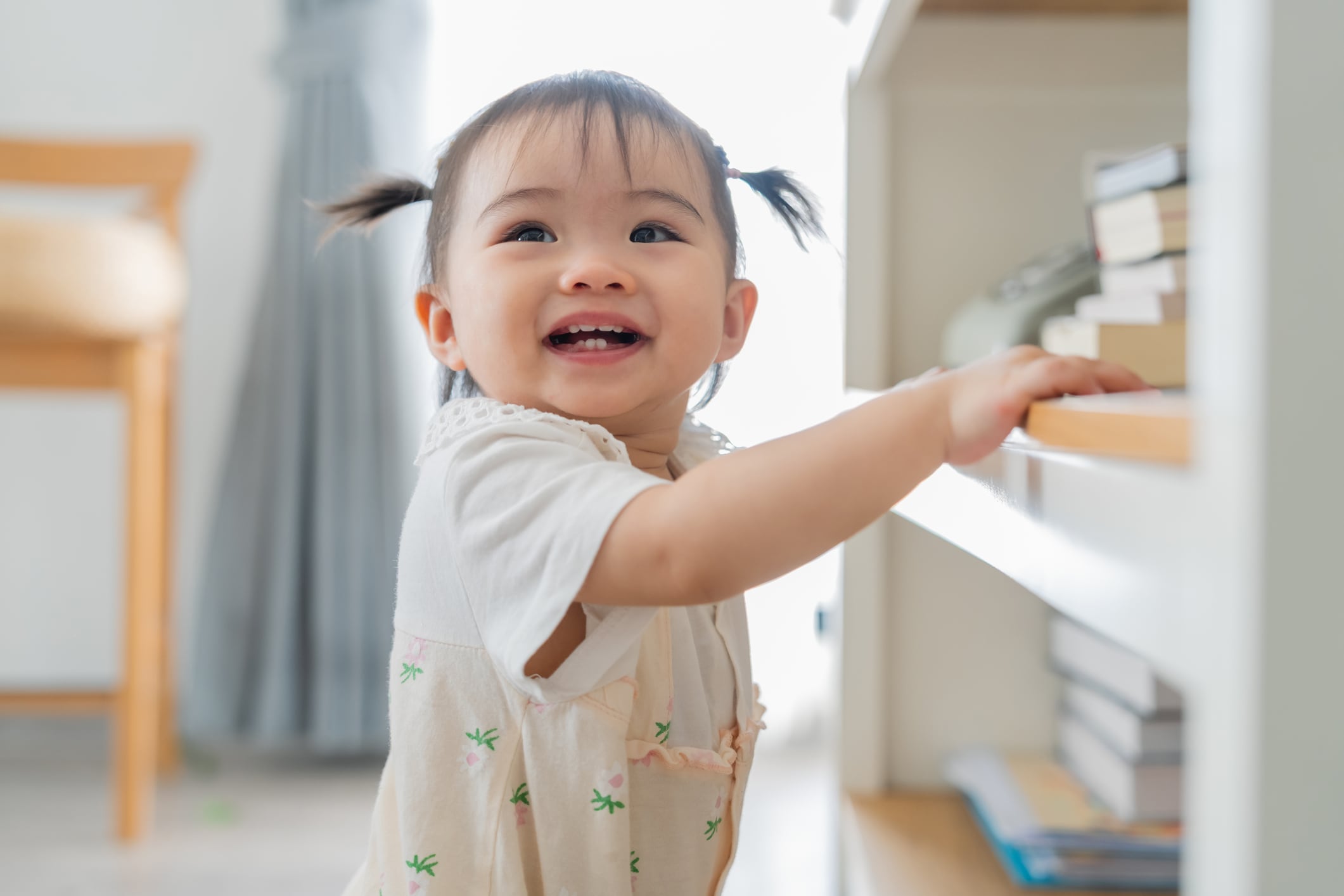 Asian baby girl learning to stand and take first steps while holding furniture at home, cute toddler exploring living room in daylight, happy childhood development moment and family lifestyle indoors.