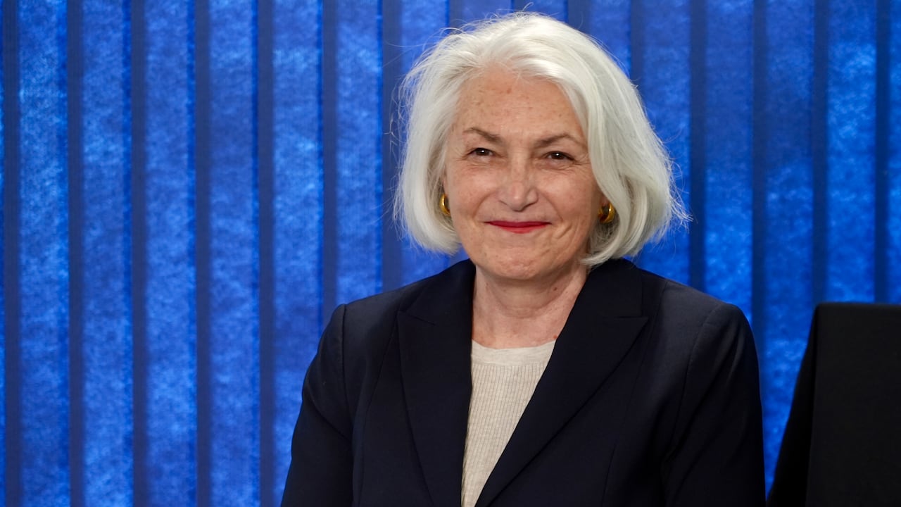 A woman with white shoulder length hair smiles in front of a blue background.