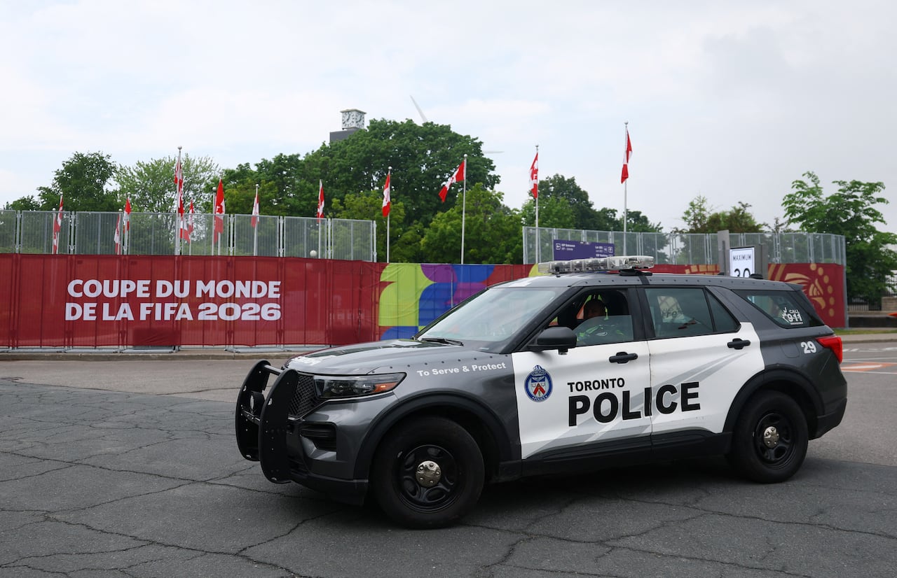 A police vehicle seen parked in a parking lot in front of a fence covered in FIFA World Cup banners and Canadian flags.