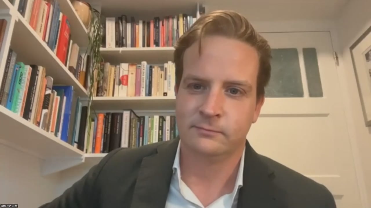 A man stares straight with books behind him.