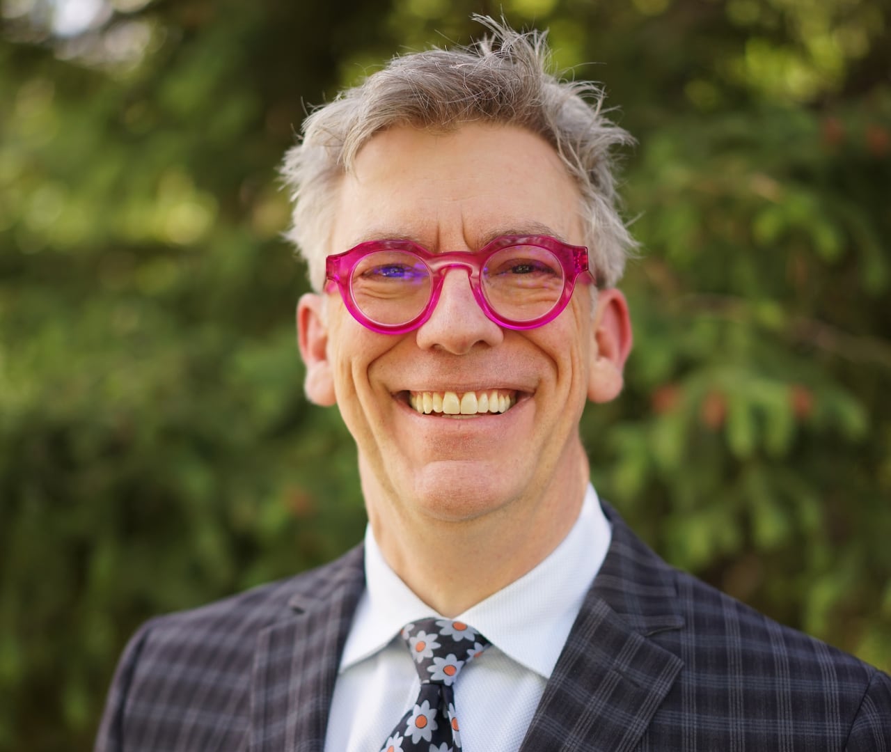 A man with grey hair and wearing pink glasses smiles at the camera.