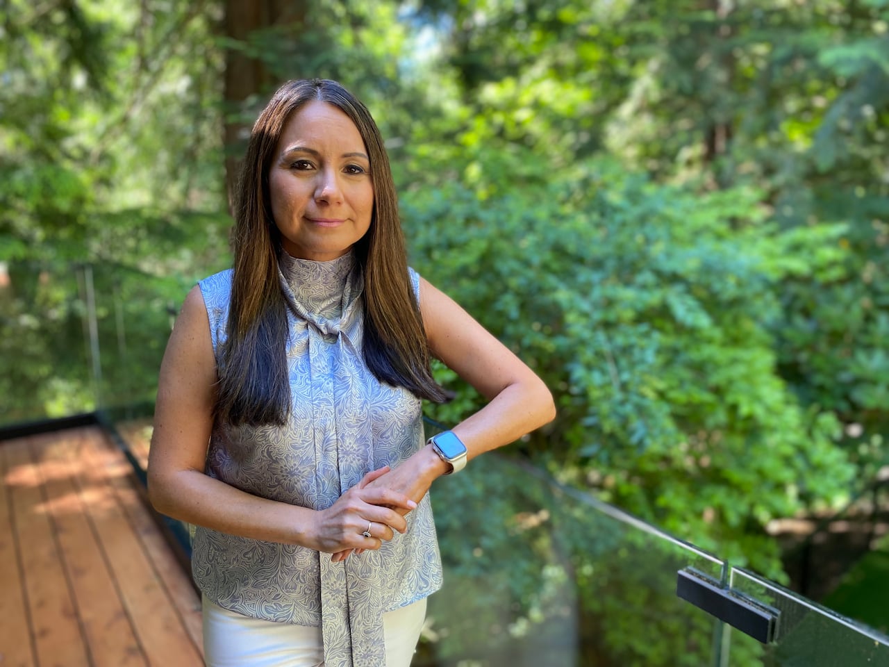 A smiling woman with long dark hair and wearing a patterned grey sleeveless blouse and white pants poses outdoors, with a leafy canopy behind her.