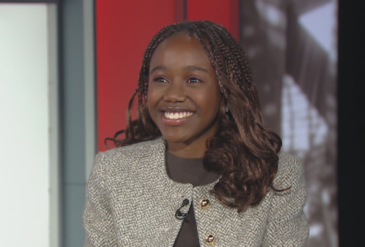 A smiling teen with long hair and a patterned beige jacket sits in a TV studio.