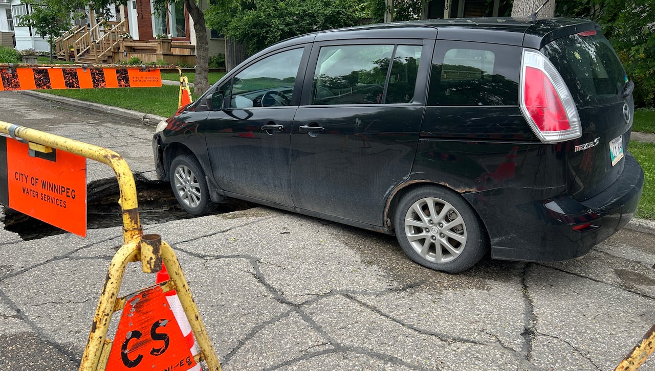 A black sedan vehicle is shown surrounded by orange pylons and construction fences after falling into a hole in a city street