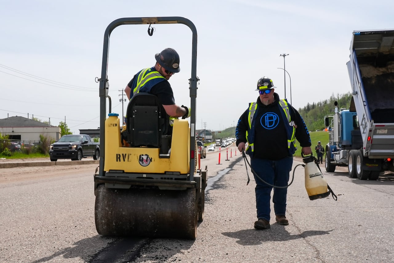 A man drives a road roller over asphalt on a busy highway as another man walks next to the vehicle.